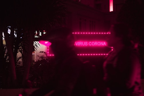 A neon pink sign displays the words 'Virus Corona' in a nighttime setting with silhouettes of people in the foreground. The environment is dark, with trees partially visible and a modern building in the background.