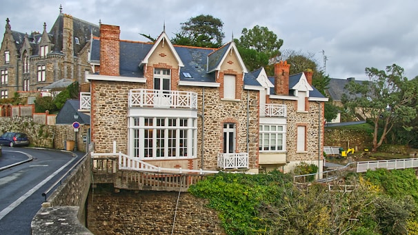 A charming stone house with white wooden balconies and red brick chimneys. It is situated on a street corner, surrounded by trees and foliage. The architecture features a mix of rustic stone walls and classic cottage-style elements. In the background, another similar building is visible, contributing to the old-world charm of the scene.