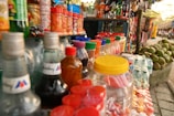 A vibrant street stall display with an array of colorful bottles and jars. The scene includes drinks in glass bottles, plastic jars filled with candies, and various snacks. Coconut fruits are stacked on the side, and vibrant textiles hang in the background.