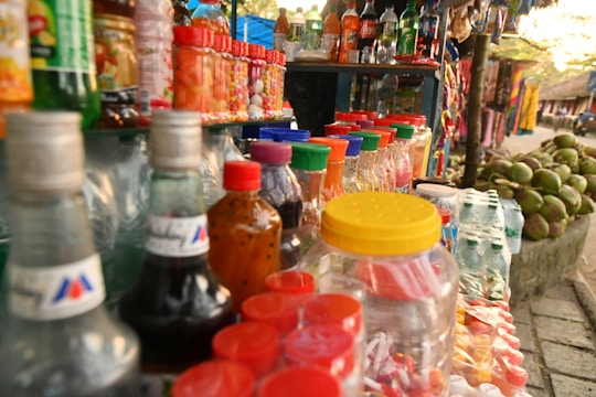 A vibrant street stall display with an array of colorful bottles and jars. The scene includes drinks in glass bottles, plastic jars filled with candies, and various snacks. Coconut fruits are stacked on the side, and vibrant textiles hang in the background.