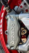 A close-up view of a silver Brembo brake caliper mounted on a vibrant red wheel. The caliper, with its brand name embossed clearly, is part of a disc brake system with visible drilled rotors and lug nuts. The metallic texture and the sleek design emphasize precision and performance.