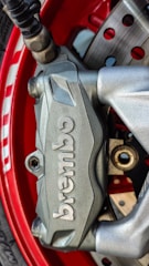 A close-up view of a silver Brembo brake caliper mounted on a vibrant red wheel. The caliper, with its brand name embossed clearly, is part of a disc brake system with visible drilled rotors and lug nuts. The metallic texture and the sleek design emphasize precision and performance.