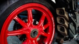 A close-up view of a motorcycle's rear wheel featuring a striking red rim. The wheel has a robust tire with visible branding, and on the right side, there are three metallic exhaust pipes. The background includes parts of the motorcycle's frame and various mechanical components.
