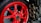 A close-up view of a motorcycle's rear wheel featuring a striking red rim. The wheel has a robust tire with visible branding, and on the right side, there are three metallic exhaust pipes. The background includes parts of the motorcycle's frame and various mechanical components.