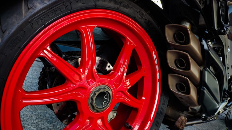 A close-up view of a motorcycle's rear wheel featuring a striking red rim. The wheel has a robust tire with visible branding, and on the right side, there are three metallic exhaust pipes. The background includes parts of the motorcycle's frame and various mechanical components.