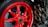 A close-up view of a motorcycle's rear wheel featuring a striking red rim. The wheel has a robust tire with visible branding, and on the right side, there are three metallic exhaust pipes. The background includes parts of the motorcycle's frame and various mechanical components.