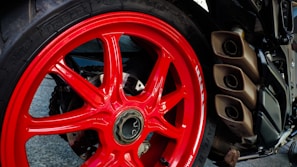 A close-up view of a motorcycle's rear wheel featuring a striking red rim. The wheel has a robust tire with visible branding, and on the right side, there are three metallic exhaust pipes. The background includes parts of the motorcycle's frame and various mechanical components.