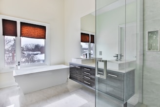 A modern bathroom with a large white freestanding bathtub positioned under two windows with brown blinds. The room features a sleek glass shower, and a countertop with dual sinks and dark wood cabinetry. Natural light fills the space, highlighting the clean and minimalist design.