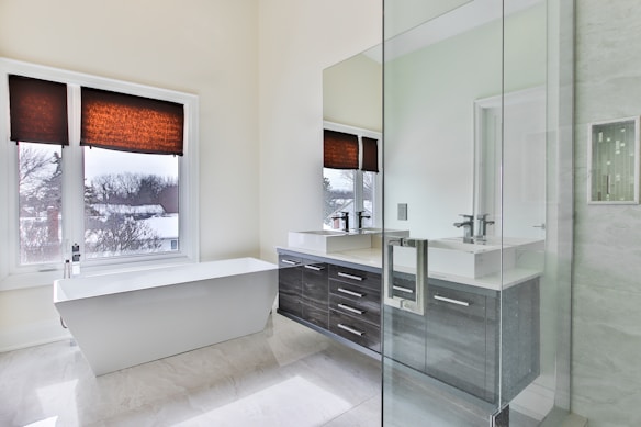 Een moderne badkamer met een groot, vrijstaand wit bad onder twee ramen met bruine jaloezieën. De ruimte beschikt over een strakke glazen douche en een aanrecht met dubbele wastafels en kastwerk van donker hout. Natuurlijk licht vult de ruimte en benadrukt het schone en minimalistische ontwerp.