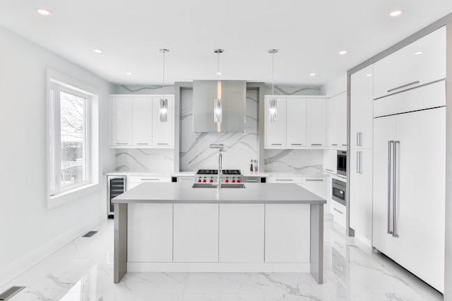 A modern, sleek kitchen featuring minimalist design with white cabinetry and marble countertops. The room includes stainless steel appliances, hanging pendant lights, and a large central island. Large windows provide ample natural light.