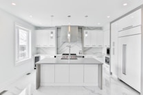 A modern, sleek kitchen featuring minimalist design with white cabinetry and marble countertops. The room includes stainless steel appliances, hanging pendant lights, and a large central island. Large windows provide ample natural light.