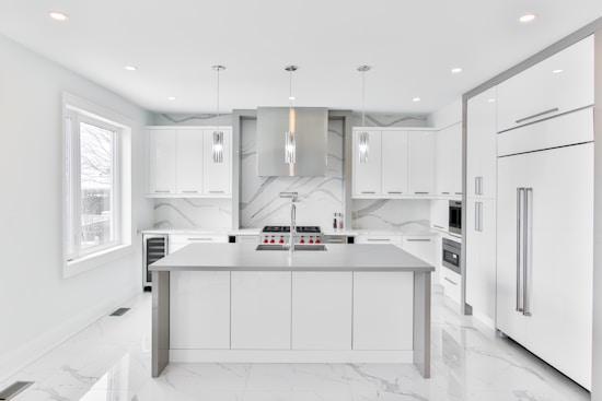 A modern, sleek kitchen featuring minimalist design with white cabinetry and marble countertops. The room includes stainless steel appliances, hanging pendant lights, and a large central island. Large windows provide ample natural light.