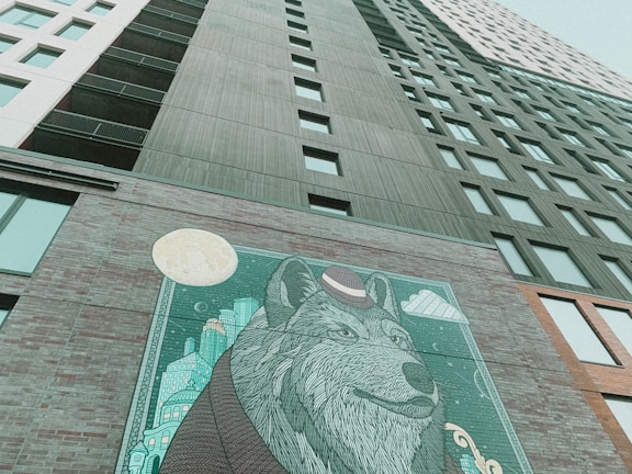 A tall modern building with numerous windows stretches upwards. On one of its lower walls is a large, artistic mural featuring a detailed illustration of a wolf wearing a hat. The backdrop of the mural includes abstract cityscapes and a prominent full moon.