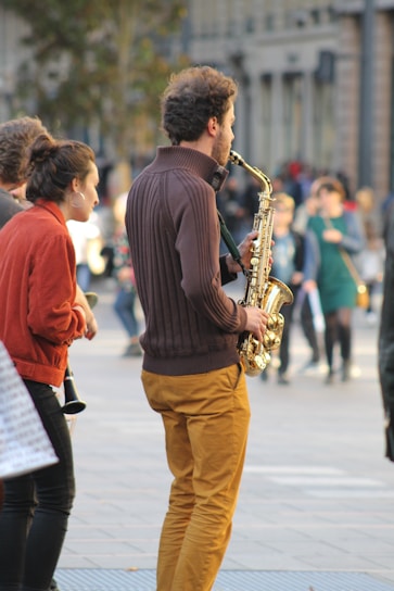 A street musician plays a saxophone while a small group of people stands nearby, enjoying the performance. The scene takes place in a spacious urban setting with blurred figures and buildings in the background, suggesting a lively atmosphere.