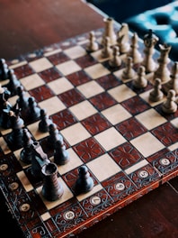 An intricately decorated chessboard with detailed, carved chess pieces set for a game. The board features a combination of light and dark squares with ornate patterns on the borders.