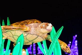 A colorful, illuminated sculpture of a sea turtle surrounded by vibrant, glowing green plants against a dark background. The turtle's shell is adorned with intricate patterns in shades of brown and tan, while the plants and background are lit in striking green and purple hues.
