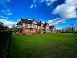 A large, historic Tudor-style house sits majestically amidst a sprawling green lawn. The structure features intricate black timber framing and brickwork, with several chimneys dotting the roof. Tall trees and well-trimmed hedges surround the house, while a vibrant blue sky with scattered white clouds stretches above.