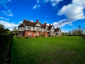 A large, historic Tudor-style house sits majestically amidst a sprawling green lawn. The structure features intricate black timber framing and brickwork, with several chimneys dotting the roof. Tall trees and well-trimmed hedges surround the house, while a vibrant blue sky with scattered white clouds stretches above.