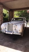 A classic white vintage Jaguar car is parked under a stone-covered carport. The car exhibits a sleek, elegant design with chrome details and multiple round headlights. Surrounded by greenery, the area is partially shaded yet illuminated by sunlight filtering through the leaves.
