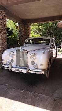 A classic white vintage Jaguar car is parked under a stone-covered carport. The car exhibits a sleek, elegant design with chrome details and multiple round headlights. Surrounded by greenery, the area is partially shaded yet illuminated by sunlight filtering through the leaves.