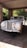 A classic white vintage Jaguar car is parked under a stone-covered carport. The car exhibits a sleek, elegant design with chrome details and multiple round headlights. Surrounded by greenery, the area is partially shaded yet illuminated by sunlight filtering through the leaves.