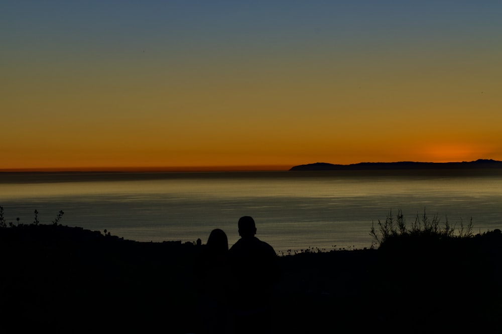 silhouette of 2 person sitting on rock near body of water during sunset