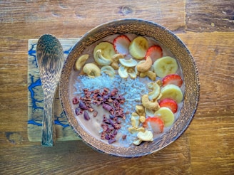 A wooden bowl filled with a smoothie topped with banana slices, strawberries, cashew nuts, shredded coconut, and cacao nibs is placed on a wooden surface. Beside the bowl, there is a wooden spoon resting on a rectangular wooden board with a blue resin inlay.