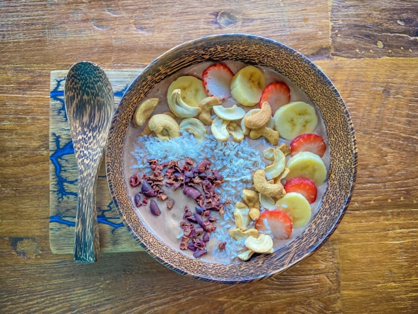 A wooden bowl filled with a smoothie topped with banana slices, strawberries, cashew nuts, shredded coconut, and cacao nibs is placed on a wooden surface. Beside the bowl, there is a wooden spoon resting on a rectangular wooden board with a blue resin inlay.
