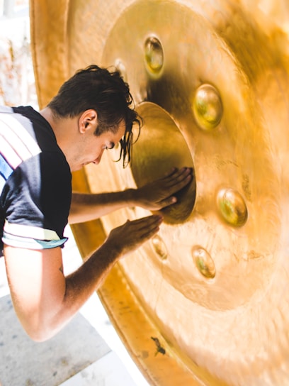 A person is leaning forward, pressing their hands against the surface of a large, shiny golden gong. The gong has several raised circular patterns and textures. The individual appears to be focused, and sunlight casts a warm glow on the metallic surface.