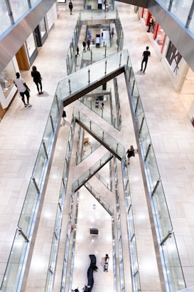 An indoor shopping mall with multiple levels visible from an overhead perspective. The design features prominent geometric patterns with angular glass railings. People are walking along the corridors, and some are visible on different floors, providing a sense of scale. The mall is well-lit with bright, neutral tones.