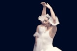 A ballet dancer is performing with arms gracefully raised above her head. She wears a white tutu adorned with delicate lace and sparkling details. Another dancer is partially visible in the background, also dressed in a similar attire. The background is dark, emphasizing the elegance and poise of the dancers.