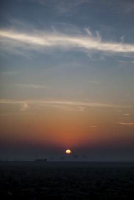 A serene landscape during sunrise with a low sun on the horizon. The sky displays a gradient of colors from dark orange near the sun to light blue at the top. Wispy clouds are scattered across the sky, enhancing the tranquil ambiance. The foreground shows a silhouette of flat terrain.