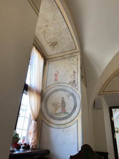 An interior view of a room featuring an ornate arched wall with faded paintings. The artwork includes circular motifs depicting a person in historical attire, engaged in an activity near flora. Light streams through a window with flowy curtains, illuminating a small table adorned with potted plants and a decorative item.