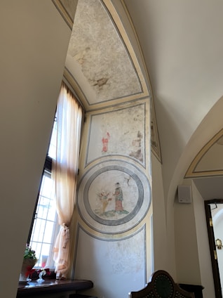 An interior view of a room featuring an ornate arched wall with faded paintings. The artwork includes circular motifs depicting a person in historical attire, engaged in an activity near flora. Light streams through a window with flowy curtains, illuminating a small table adorned with potted plants and a decorative item.