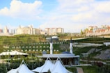 A large, elegant building resembling a palace is situated on a lush, green hillside surrounded by smaller buildings and apartments. A mosque with a prominent dome and tall minarets is in the foreground. The area is landscaped with expansive lawns, pathways, and neatly arranged trees. The sky is bright with scattered clouds, adding a serene atmosphere.