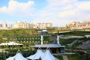 A large, elegant building resembling a palace is situated on a lush, green hillside surrounded by smaller buildings and apartments. A mosque with a prominent dome and tall minarets is in the foreground. The area is landscaped with expansive lawns, pathways, and neatly arranged trees. The sky is bright with scattered clouds, adding a serene atmosphere.