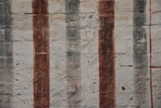 Vertical stripes of varying shades and textures are painted over a rough, textured wall. The surface shows signs of wear and age, with muted tones enhancing its rustic appearance.