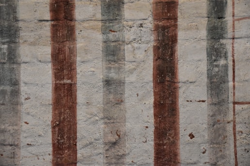 Vertical stripes of varying shades and textures are painted over a rough, textured wall. The surface shows signs of wear and age, with muted tones enhancing its rustic appearance.
