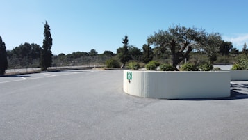 The image features an empty parking lot surrounded by trees and a low concrete barrier. The trees and vegetation around the lot suggest a natural setting. The sky is clear and blue, indicating a sunny day.