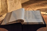 An open book displayed on a stand with visible text written in a Gothic script. The background consists of a rough, textured wall and a brick floor, suggesting an old or historical setting.