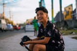 A young teenager smiling brightly while holding a camera in a vibrant urban street.