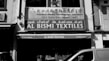A black and white photo of a storefront with signs in multiple languages. The main sign reads 'AL BISMI TEXTILES' located at 122 Dunlop Street, Singapore, with a phone number provided. Above it, there is another sign for the 'Singapore Noodles Manufacturers Association.' In front of the store, various textiles are displayed, and a partial view of a van is visible.