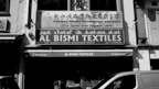 A black and white photo of a storefront with signs in multiple languages. The main sign reads 'AL BISMI TEXTILES' located at 122 Dunlop Street, Singapore, with a phone number provided. Above it, there is another sign for the 'Singapore Noodles Manufacturers Association.' In front of the store, various textiles are displayed, and a partial view of a van is visible.
