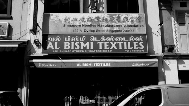 A black and white photo of a storefront with signs in multiple languages. The main sign reads 'AL BISMI TEXTILES' located at 122 Dunlop Street, Singapore, with a phone number provided. Above it, there is another sign for the 'Singapore Noodles Manufacturers Association.' In front of the store, various textiles are displayed, and a partial view of a van is visible.