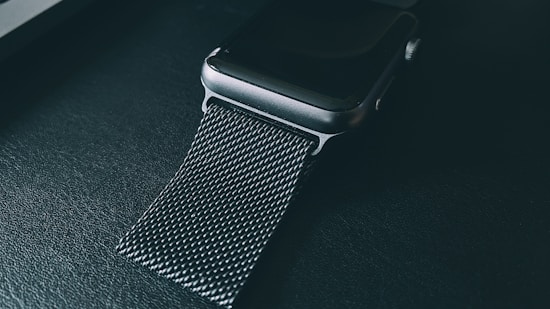A close-up view of a smartwatch with a dark, metallic mesh band resting on a textured surface. The lighting is low, highlighting the sleek design and detail of the watch strap.