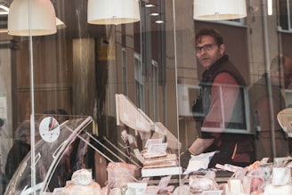 A man standing behind a counter inside a shop, presumably a delicatessen or bakery, filled with various food items such as cheeses and meats on display. The shop's interior has a warm, cozy ambiance, emphasized by hanging lights. The man is looking directly ahead while the shop window has reflections of the street and possibly other people passing by.