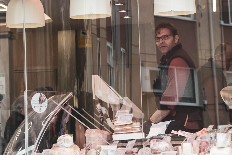 A man standing behind a counter inside a shop, presumably a delicatessen or bakery, filled with various food items such as cheeses and meats on display. The shop's interior has a warm, cozy ambiance, emphasized by hanging lights. The man is looking directly ahead while the shop window has reflections of the street and possibly other people passing by.