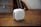 A compact white portable speaker with a perforated front grill and a leather strap is placed on a wooden surface. In the background, parts of a shelf or furniture and books are visible, creating a cozy and minimalistic ambiance.