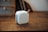 A compact white portable speaker with a perforated front grill and a leather strap is placed on a wooden surface. In the background, parts of a shelf or furniture and books are visible, creating a cozy and minimalistic ambiance.