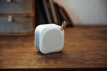 A compact white portable speaker with a perforated front grill and a leather strap is placed on a wooden surface. In the background, parts of a shelf or furniture and books are visible, creating a cozy and minimalistic ambiance.